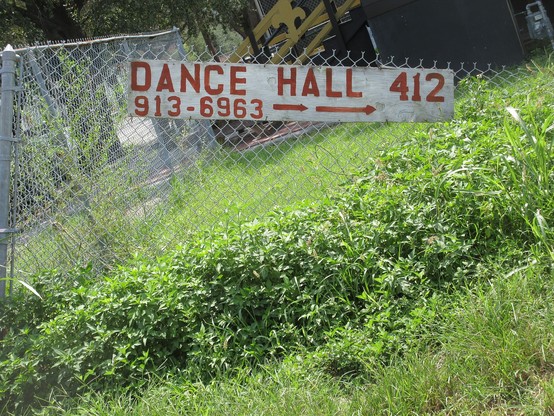Outdoor scene, chain link fence partly overgrown with weeds. A faded wooden sign with red letters on white background is attached to the fence. It has an arrow pointing right. Text: "DANCE HALL 412" and a 7 digit telephone number. 