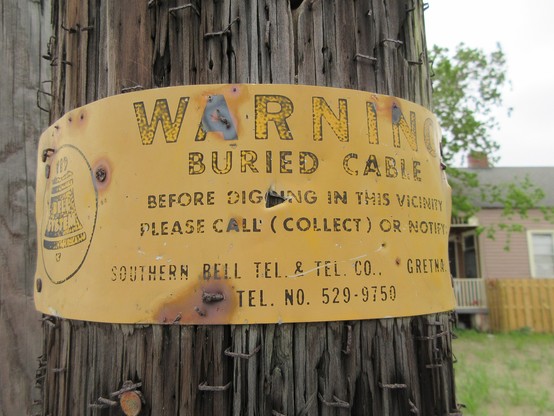 Wooden utility pole with a faded metal sign attached, black letters on a yellow background. At right is artwork depicting a stylized bell, labeled "BELL SYSTEM".   Text: "WARNING - BURIED CABLE - BEFORE DIGGING IN THIS VICINITY PLEASE CALL (COLLECT) OR NOTIFY - SOUTHERN BELL TEL & TEL CO., GRETNA - TEL. NO." (7 digit telephone number) 
