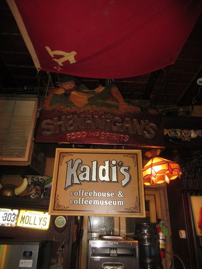 Interior of part of space under ceiling of French Quarter bar with many old momentos and decorations hanging.  Seen are old stained glass lamps, license plates, and shop signs.  Centered is a sign reading "Kaldi's coffeehouse & coffeemuseum". 
