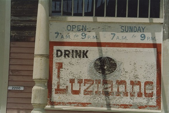 Faded wooden sign built into wall of old wooden building.  Top of sign text "OPEN 7a.m. to 9p.m. - SUNDAY 7a.m. to 9p.m."
Below artwork depicting a cup of coffee, with larger text "DRINK LUZIANNE"