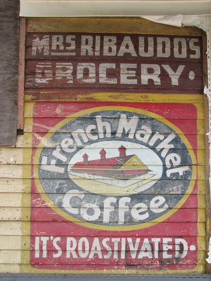Old shop signage painted on to weatherboards of wooden building.  Text at top: MRS. RIBADUO'S GROCERY. "
Below is the logo for French Market brand coffee, art in the center depicts a single story building with high roof and three ventilation towers - older building of the Old French Market in New Orleans.  In an oval around the artwork text reads "French Market Coffee".
Text below: "IT'S ROASTIVATED."
