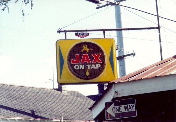 Outdoor sign hanging from metal frame off single story corner building.  Logo of a man on horseback on tap (depicting the Andrew Jackson statue in Jackson Square, New Orleans, the trademark of Jax beer) and a stylized beer tap. Text: "JAX - ON TAP". 
