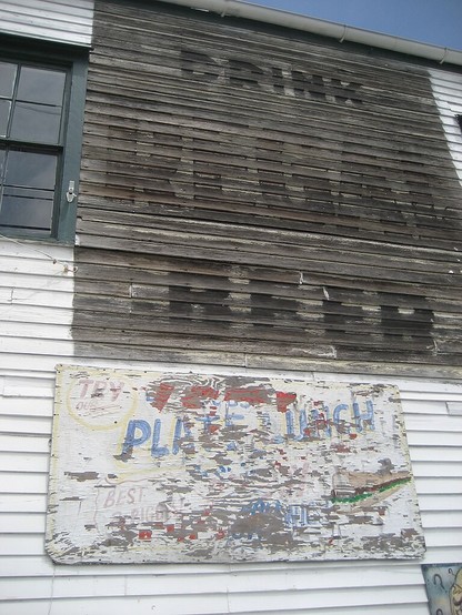Very faded large painted letters on weatherboards of 2 story wooden building, reading "DRINK REGAL BEER". 

Another faded wooden sign is below, almost all illegible other than the phrase "PLATE LUNCH". 