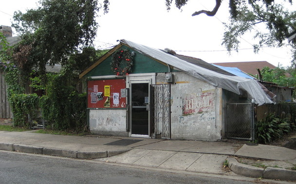 A small neighborhood bar in a decrepit looking building with tarped roof.  Glass and metal door at center, above which is an old Christmas wreath.  To left of door is a section of wood with various posters and flyers stapled to it (details not legible). To right side of door is faded painted sign "REGAL". 
