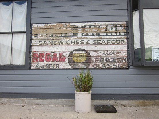 Section of exterior wall of wooden building, with weatherboards recently repainted except for a rectangle painted with faded advertising. 
Text: "DIXIE TAVERN - SANDWICHES & SEAFOOD". 
Below artwork depicting a glass of beer. 
TEXT: "REGAL keg BEER - With FROZEN GLASSES". 
