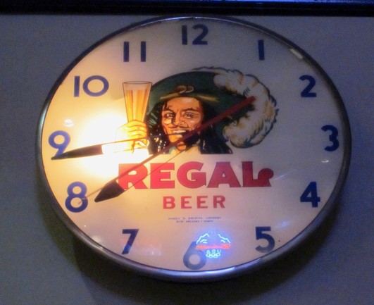 Illuminated analog clock on wall.  In center is artwork of the face and hand of a man holding a glass of beer. The man has long black hair and a pencil moustache, and is smiling with enthusiasm.  He wears a 17th century style soft broad-brimmed hat with a large plume, like a Musketeer. 

Text below: "REGAL BEER". 

