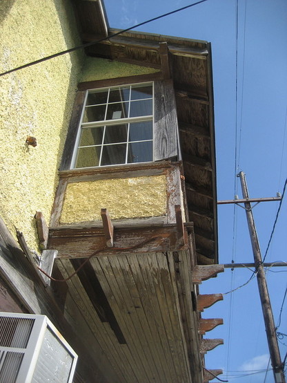 Detail of wood and stucco building, a protruding balcony window upstairs; some of the wooden slats below the window have decayed and fallen down. 