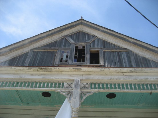 Detail of 19th century wooden building with some carved "gingerbread" style decorative wooden details.  Looking to set of three windows in center of attic above porch; most of the panes of glass are missing. 