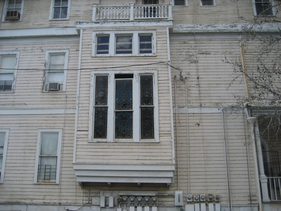 Side of a once grand old  3 story house divided into apartments. Some windows have air-conditioners, some drawn shades, and some wooden boards behind them. At center a large window retains old decorative stained glass; the top of the center pane is slightly open. 