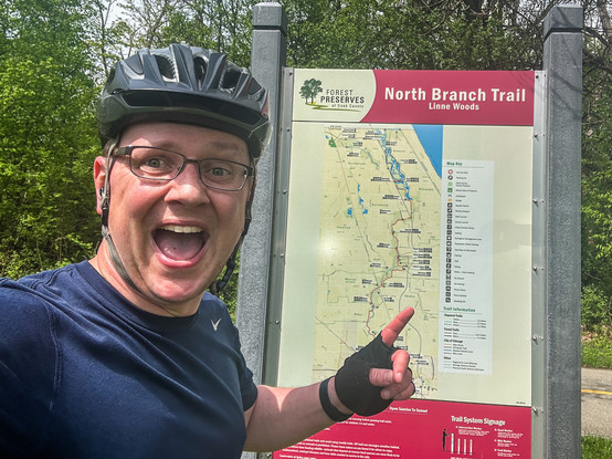Person in cycling gear taking a selfie with a smile, pointing at a map sign for the North Branch Trail Line Woods.