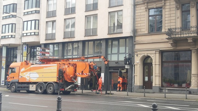 Un camión color naranja con la inscripción Kanalreinigung parado y ejecutando la actividad que se describe en el toot, con dos operarios apoyados en la fachada del edificio, observando el paisaje mientras se fuman un pitillo.