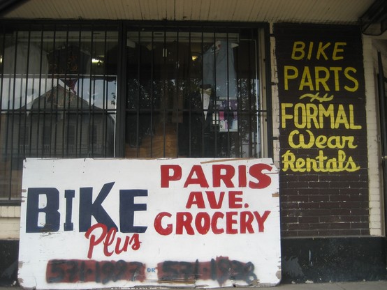 Urban streetscape, glass windows with bars, hand painted signs. 

Painted on wooden weatherboards of building at right, text: "BIKE PARTS - FORMAL Wear Rentals". 

Below, wooden sign on sidewalk leaning against building, text: "BIKE Plus - PARIS AVE. GROCERY", telephone number at bottom mostly obscured with painted over blotches. 