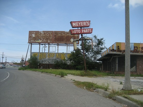 Commercial building that looks long deserted at curve in a road.  

Billboard type signs beside the building; the upper signs have faded to brown with nothing legible. The bottom sign is less faded, with text: "HUB CAP KING - AUTO PARTS".  

Sign on metal pole text: "MEYER'S - AUTO PARTS". 