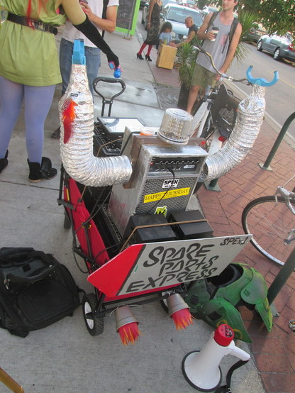 A miniature parade float made out of a meter long pull wagon is on a sidewalk.  Sign at back reads "SPARE PARTS EXPRESS". 
In the cart is an audio speaker decorated with pipes, plastic clips, and other inexpensive items made to resemble a 1950s science fiction show robot.  Two plastic cups are attached at bottom back of the wagon with red and orange paper streamers, to depict rocket engines firing. 