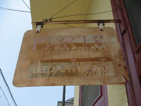 Faded metal sign hanging on pole from building.  Two or three different layers of painted text are partially visible in overlayed faded letters.  The most clearly readable parts of sign are block letters at top, text: "PARTS".  Lower section of sign in different cursive text: "Welcome Here". 
