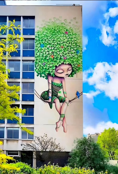 Streetartwall. This charming comic-style mural of a girl is sprayed/painted on the exterior wall of a seven-story hospital building. The girl is wearing a green dress made of woven vines and leaves. She has large eyes, a small nose and a small mouth. Her fuzzy hair consists of green, heart-shaped leaves that look like a tree crown and also contain small, colorful flowers. She is sitting on a swing made of two tendrils and is looking at a small blue bird sitting next to her. An enchanting spring mural in a children's hospital.