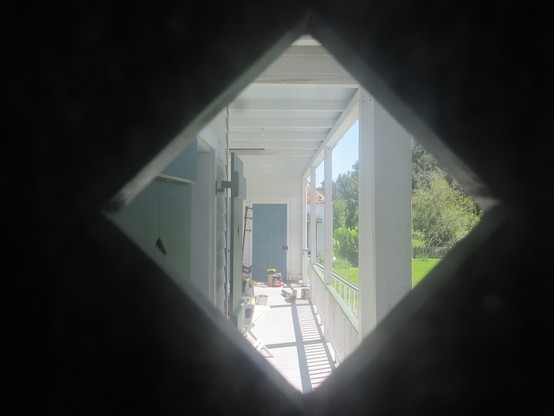 View out through square window set with points at top bottom and sides, diamond shape.  Through window is a long narrow roofed porch of a wooden building. Along the left side and in distance straight ahead are wooden shutters covering windows and doors. On the porch are various small items, including cans of paint, a bucket, small folding tables, a leaf blower, and a bag of charcoal.
At right, stairs lead down about a meter to the grounds.   Greenery of lawn, small trees and large leafed plants…