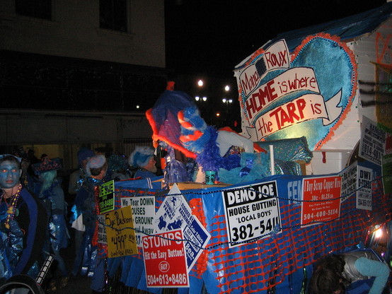 Small parade float, raised section with large letters reading: "Mama Roux - HOME is where the TARP is".  The float is decorated with blue FEMA tarps and multiple plastic signs as common in the months of slow recovery after the Hurricane Katrina Federal Flood disaster in New Orleans, advertising services with telephone numbers.  Various sign texts seen: "DEMO JOE - House Gutting - Local Boys". "I Buy Scrap Copper Wire".  "We Tear Down Houses to the Ground".  "We Sue".  "Flooded by Katrina - Scre…