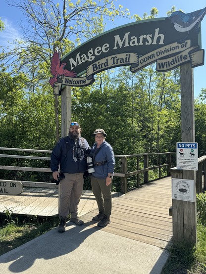 A man and a woman stand side by side under a sign reading “Magee Marsh Bird Trail” with figures of a Northern Cardinal and Black-throated Blue Warbler on the sign. They are both wearing lots of camera gear and binoculars, and are dressed for the outdoors. It’s a beautiful sunny day with the blue sky over trees and shrubs full of fresh green leaves