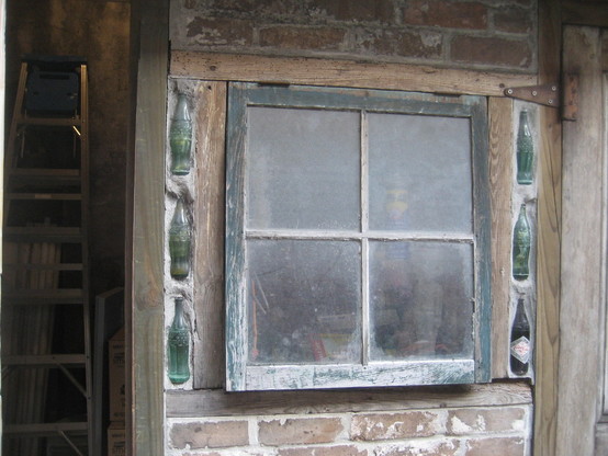 Detail part of brick building showing window and what looks like an informal mid-20th century renovation of a much older building. 

4 pane square window with hinges on top, with wooden beam frames, in brick wall.  Space to right and left of window framing is filled in with concrete, into which are three old glass Coca-Cola bottles on each side, facing upwards in vertical rows. 