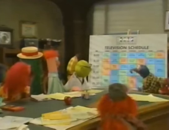 Puppet characters gathered around a table looking at a large television schedule chart.