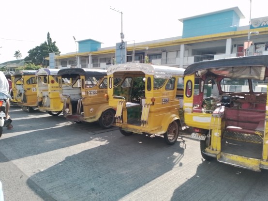 Several yellow tricycles parked perpendicular to road in front of a public market.