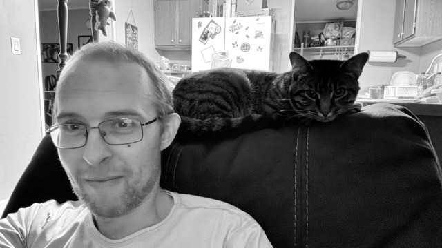 A white guy with glasses taking a selfie on a couch with a cat lying across the back. Both are looking at the camera. My neighbors messy kitchen in the background. 