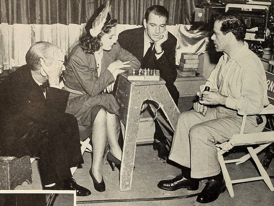 Barbara Stanwyck and Frank Capra play chess on a portable board set up on a sawhorse while Gary Cooper watches.