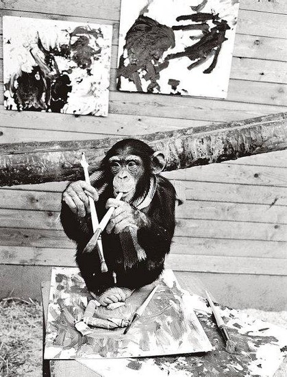 Das Schwarz-Weiß Foto zeigt einen Schimpansen mit Pinseln in den Händen. Im Hintergrund sind Gemälde zu sehen
