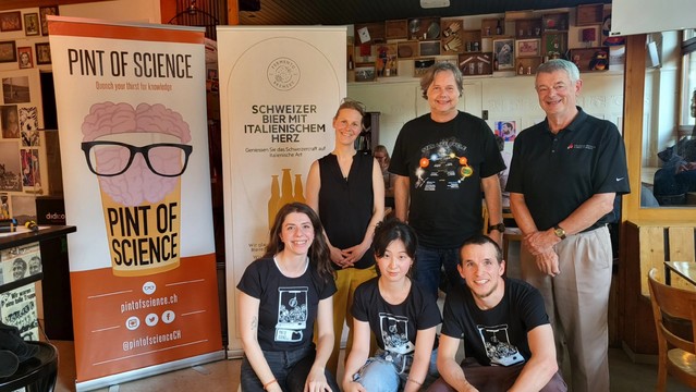 group photo of three speakers standing in the back with three organisers sitting in front of them beside a banner with a Pint of Science logo