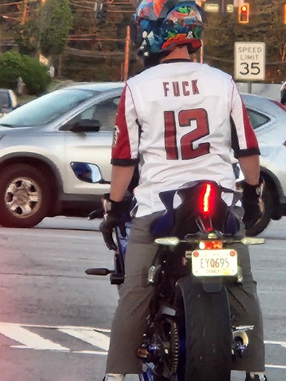 A motorcycle rider at a stoplight. Wearing a sports jersey that says fuck 12