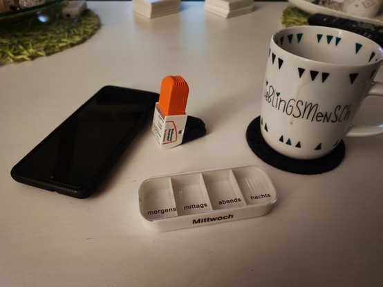 Asthma Spray, leere Tablettendose für Mittwoch, ein Handy und eine große Tasse mit Kaffee und dem Schriftzug 