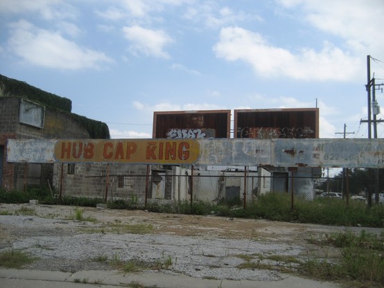 Faded paint on metal signage behind empty parking lot with spots of weeds growing.  Text in large red letters on yellow background: "HUB CAP KING".  The "K" letter is decorated with a crown at top. 