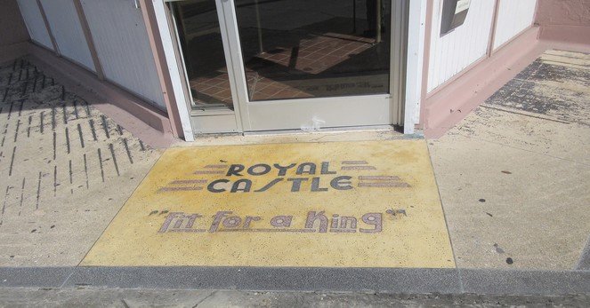 Signage embedded in concrete in front of corner door entrance. 
Text: "ROYAL CASTLE - fit for a King". 
