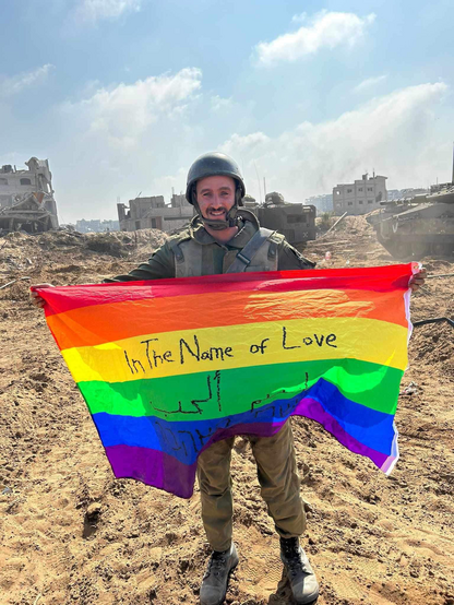 An image of an IDF soldier raising the rainbow flag in front of ruins in Gaza. On the flag the soldier has written “in the name of love.”