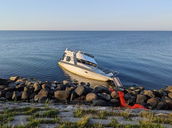 Photo of a shipwrecked motorized boat. It is about a quarter under water, lying near some big rocks at the shoreline. In the background is the ocean - looking very still.