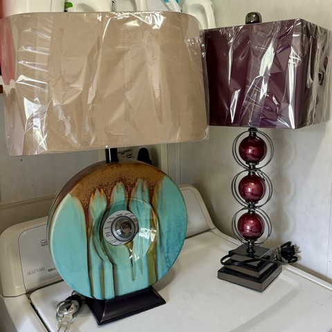Two table lamps on a Maytag washing Machine. The lamp to the left is a round, ceramic torus for a body that is glazed in a light blue and rusty brown colors on a black, square base with a rounded rectangular shade. The Lamp to the right’s body is comprised of three red spheres stacked atop one another within individual rings and with a rectangular red shade on a dark gray metallic pedestal base.