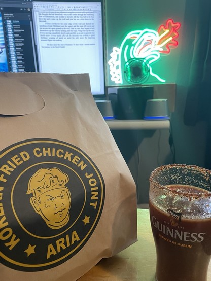 A bag of Korean fried chicken, a michelada with a tajin rim, my computer monitor, and my neon Godzilla sign.