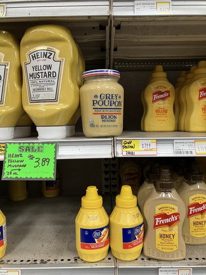 Display of several brands of yellow mustard on a grocery store shelf, all with narrow squirt caps. In the middle is a wide top jar of honey mustard.