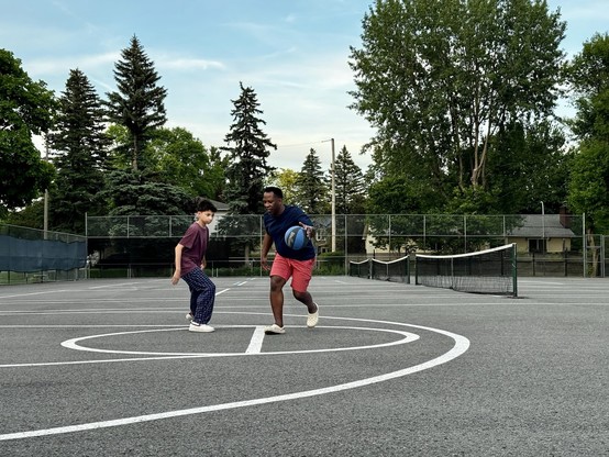 Two people playing basketball on an outdoor court, surrounded by trees and a tennis court in the background.