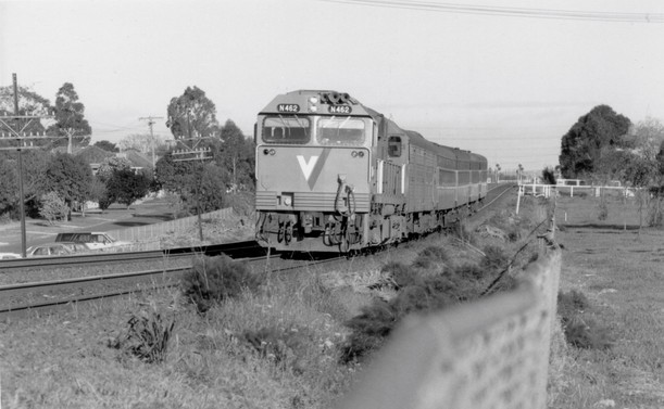 A black and white photo taken looking over a lineside cyclone wire fence of a diesel loco-hauled train heading towards us along a double track mainline railway.  The train, which has a VLine N class loco on the front, comprises five or six carriages and is traversing a dip in the railway with the loco heading slightly uphill whilst the rear car seems to still be headed downhill.
