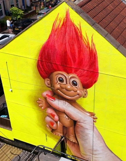 Streetartwall. A mural of a toy troll was sprayed/painted on a somewhat hidden exterior wall from the first floor to the roof gable. The background is bright yellow. A hand with red-painted fingernails holds a plastic doll with upright, long red hair. A grinning troll that became very popular in the 70s.
Info: The troll doll or magic troll (Hasbro) was invented by the Danish woodcarver Thomas Dam (1915-1989) and collected several times over the decades. There are now also cartoon series and films.