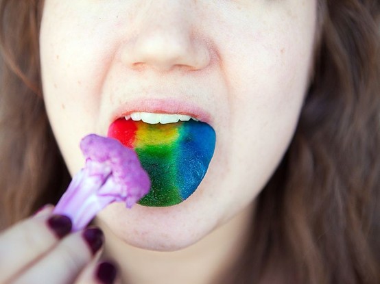 Une personne à la langue peinte aux couleurs de l'arc-en-ciel fait mine de manger un broccoli mauve