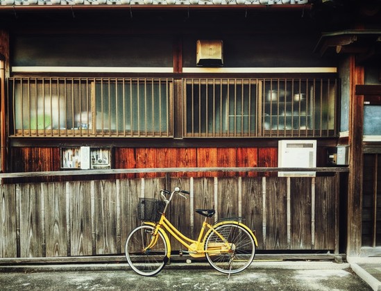 A yellow bicycle standing next to a wooden railing and wood siding of a building. The wood above the railing is vibrant brown and is faded below. Glass sliding windows with vertical metal bars are above the wood and along the whole side of the building