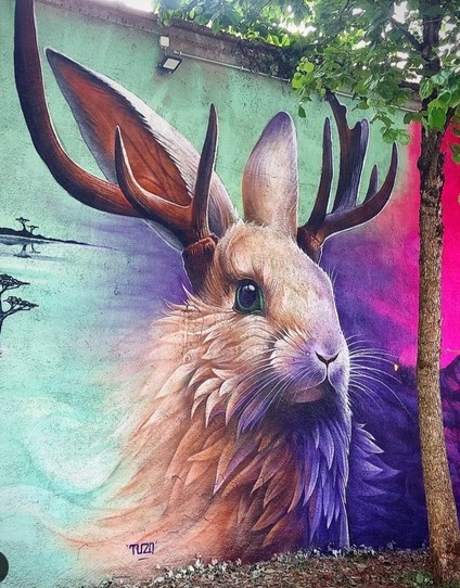 Streetartwall. A mural with a funny mythical creature called "Wolpertinger" (from Germany, Bavaria) was sprayed on a street wall. The creature is a rabbit with deer antlers and fluffy beige fur. The background is a bright purple and green and shows a minimalist depiction of a river landscape.
Info: The Wolpertinger is a Bavarian mythical creature whose exact origin is unclear. At the beginning of the 19th century, taxidermists probably assembled new animals from parts of different animals and sold them to tourists. It can therefore have different shapes, for example a squirrel with a duck's beak or a hare with duck wings...
