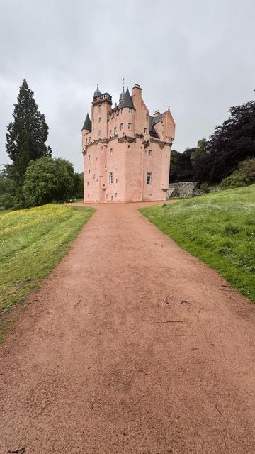 A pink-colored castle with multiple turrets is situated at the end of a gravel path, surrounded by greenery under a cloudy sky.