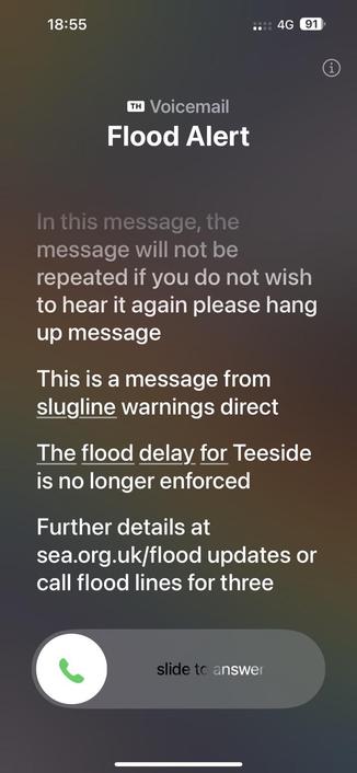 Screenshot of a mobile voicemail notification about a flood alert. The message indicates that the flood delay for Teesside is no longer enforced and includes a website for further details. The phone shows the option to slide to answer the call.