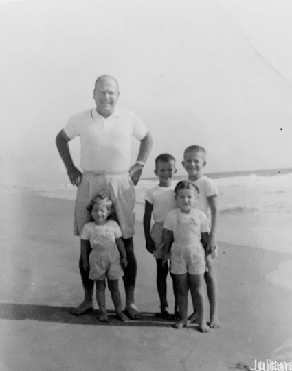 A black and white Brownie box camera image of a sturdy man on a beach with his hands on his hips and four young children clustered around his legs. All are wearing shorts and white shirts. Two siblings are yet to be born.