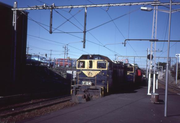 A photo taken from a passenger platform at a railway station of a blue and yellow liveried Y class diesel shunting locomotive hauling a passenger train of red-liveried wooden bodied passenger carriages towards us and into the station.