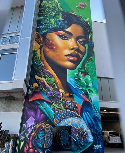 Streetartwall. The imaginative mural of a mysterious young woman is sprayed/painted on the concrete wall of a modern three-storey building. The head of a colored woman with black hair is depicted in the middle. She is looking down at the viewer from above. The rest of the wall is richly detailed and colorful. A blue fish with orange lines swims directly below the woman.Otherwise, she is surrounded by abstract wavy and geometric patterns in purple, orange and green. A small colored cube can be seen at the top right. A wild mixture of pop art and fantasy manga, but united in a flowing portrait.